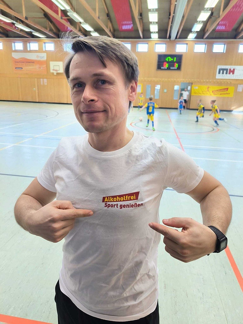 Trainer zeigt stolz auf den Schriftzug von "Alkoholfrei Sport geniessen" auf seinem weißen T-Shirt.