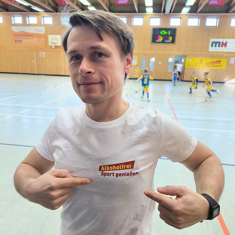 Trainer zeigt stolz auf den Schriftzug „Alkoholfrei Sport geniessen“ auf seinem weißen T-Shirt.